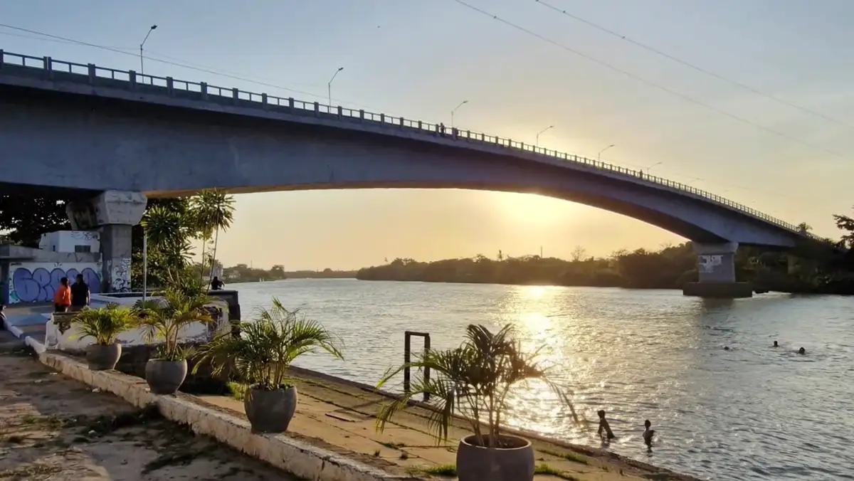 Pôr do sol sob o arco da ponte sobre o rio Igaraçu, em Parnaíba, visto da orla de pedra com palmeiras em vasos